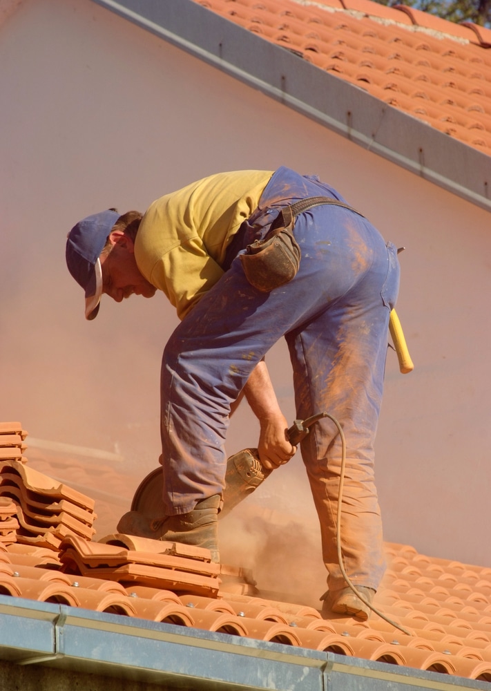 Couvreur professionnel coupe de tuiles sur le toit Couvreur concentré sur le toit, vêtu de bleu, coupant des tuiles en terre cuite avec une meuleuse, entouré d'un nuage de poussière orange.