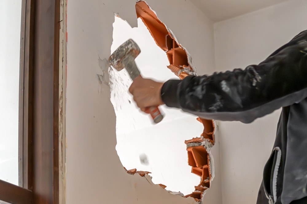 Démolition de mur au marteau pour travaux de rénovation. Démolition d'un mur intérieur avec un marteau. Le coup brise le plâtre, exposant la structure en brique orange et projetant des débris.