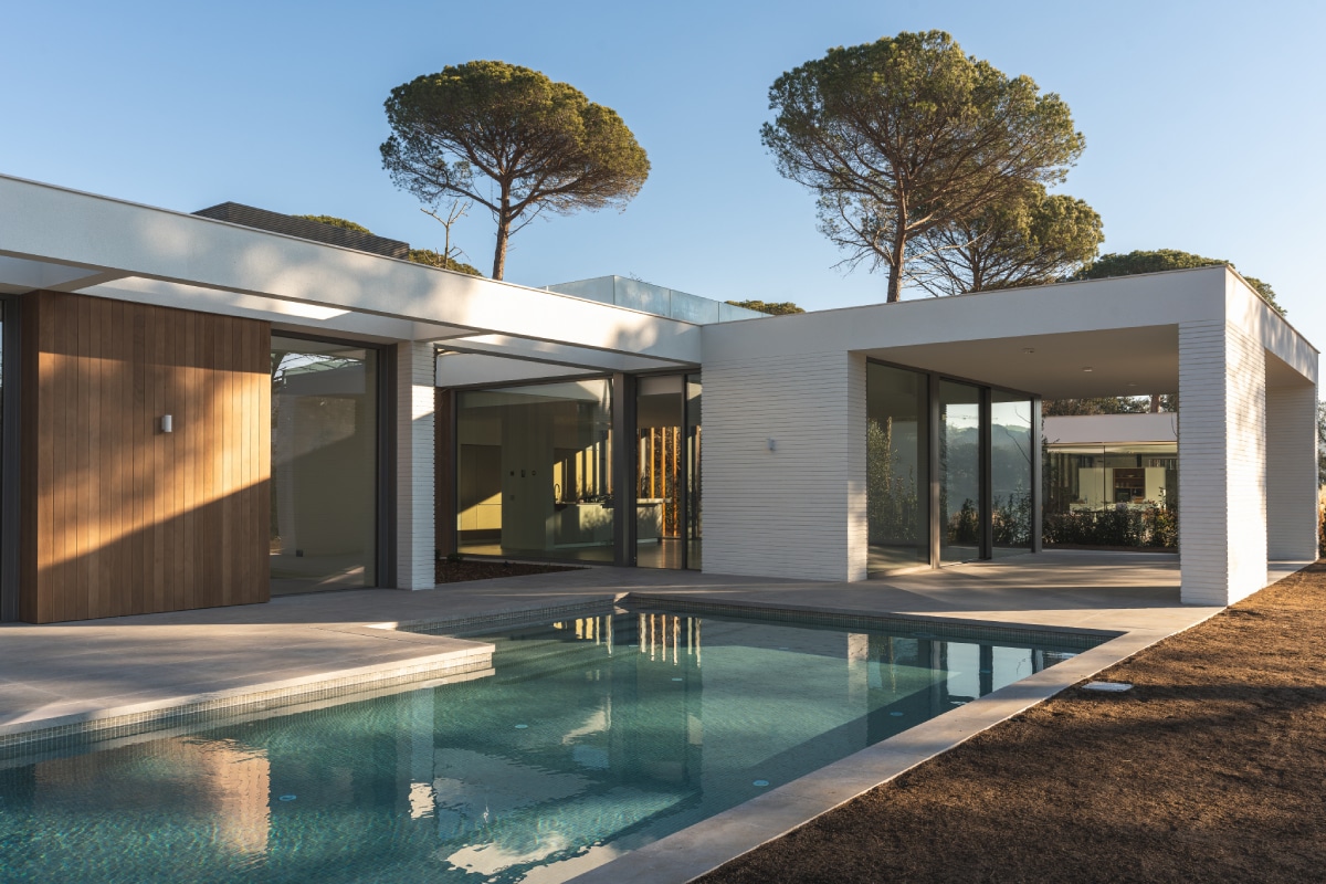 Architecture moderne et piscine : Villa de luxe sous les pins. Maison moderne à toit plat avec piscine turquoise, façades blanches et vitrées. Deux grands pins parasols dominent la structure.