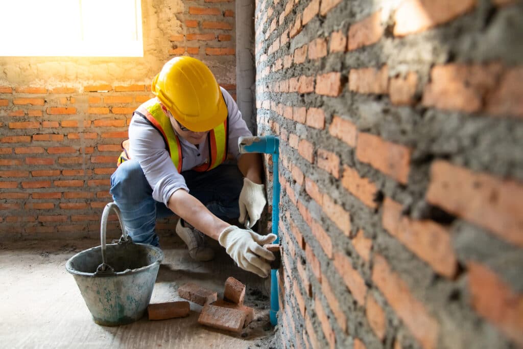 Chantier : Pose de tuyau PVC sur mur de briques par un ouvrier. Ingénieur ou maçon en casque jaune travaillant sur un tuyau PVC fixé à un mur de briques. Inspection de la plomberie sur chantier.