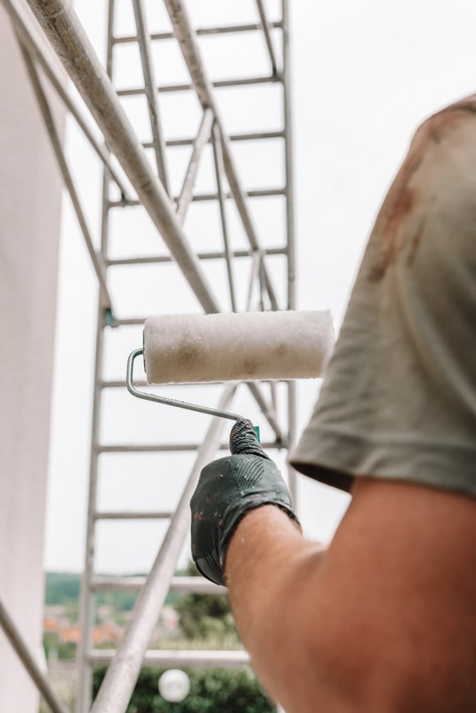 Peinture façade : travaux de rénovation sur échafaudage Ouvrier en gant noir peignant une façade à l'aide d'un rouleau sur un échafaudage. Travaux de rénovation extérieure sous ciel clair.