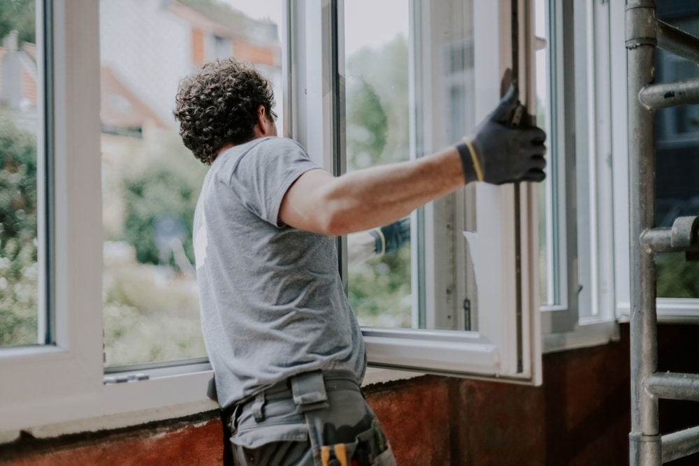 Ouvrier ajuste un châssis de fenêtre PVC blanche. Rénovation de façade avec travaux de menuiserie et vue sur la nature.