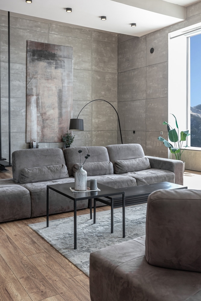 Salon moderne : Canapé gris modulaire et mur en béton minimaliste Intérieur moderne : canapé modulaire gris, sol en bois, tables noires et mur en béton texturé. Grande fenêtre avec vue montagne.