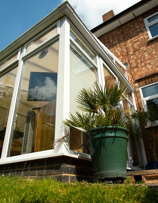 Véranda blanche moderne pour maison en briques et jardin. Véranda blanche moderne adossée à une façade en briques sous un ciel bleu. Un palmier en pot vert sur l'herbe au premier plan.